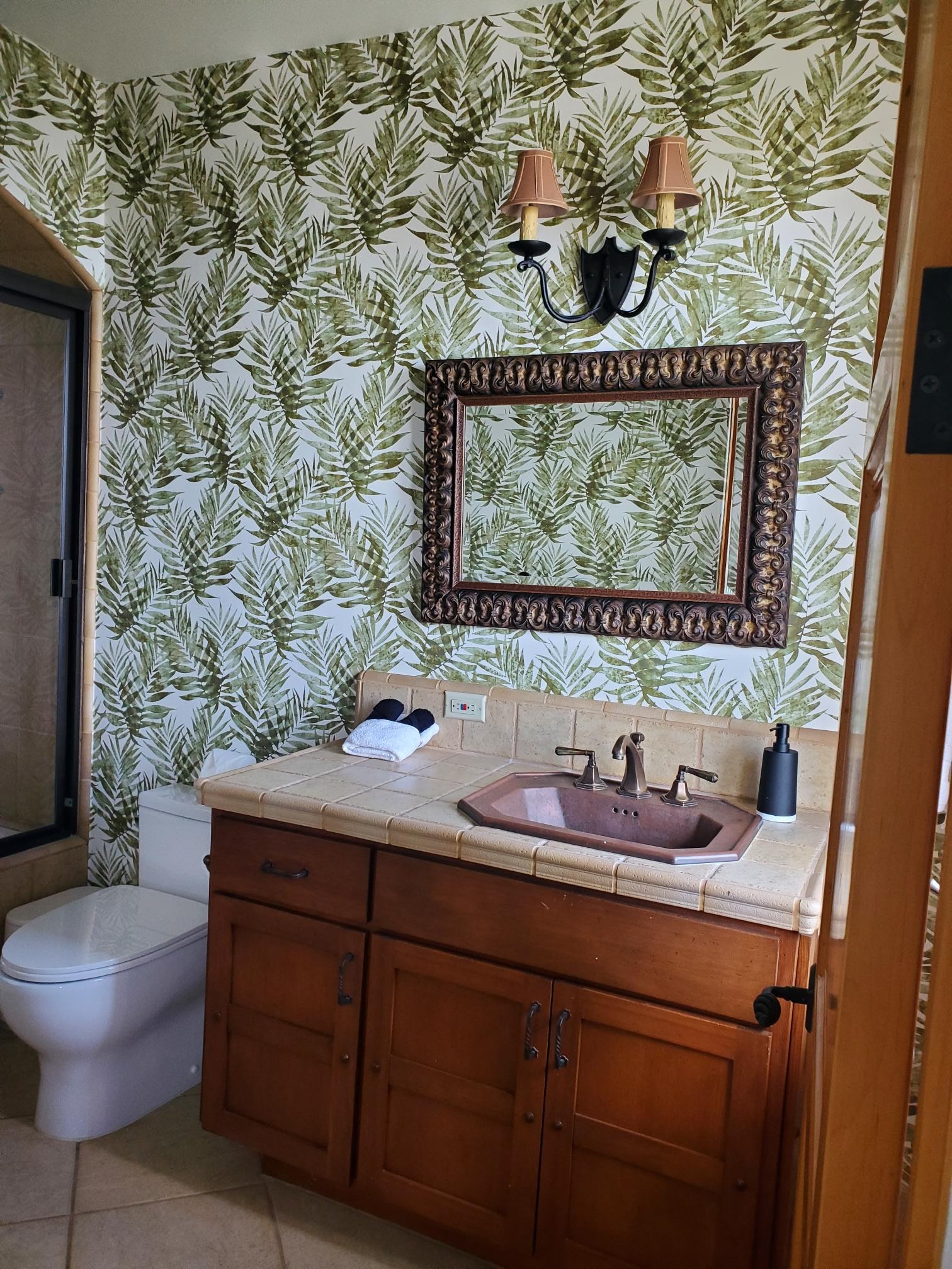 Powder room with botanical wallpaper, carved mirror, and warm wood vanity