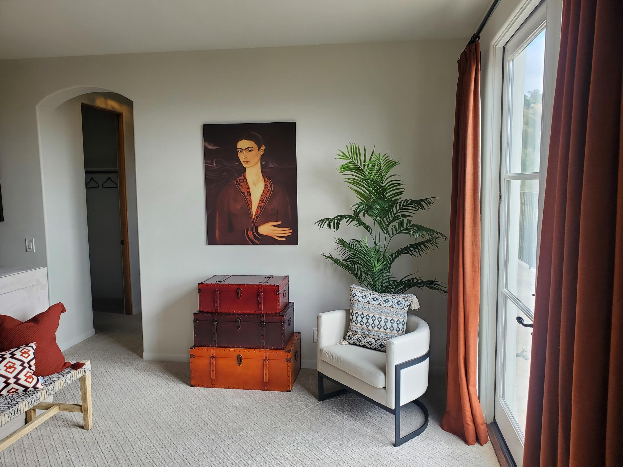 Sitting area with stacked vintage trunks, white chair, palm, and rust curtains